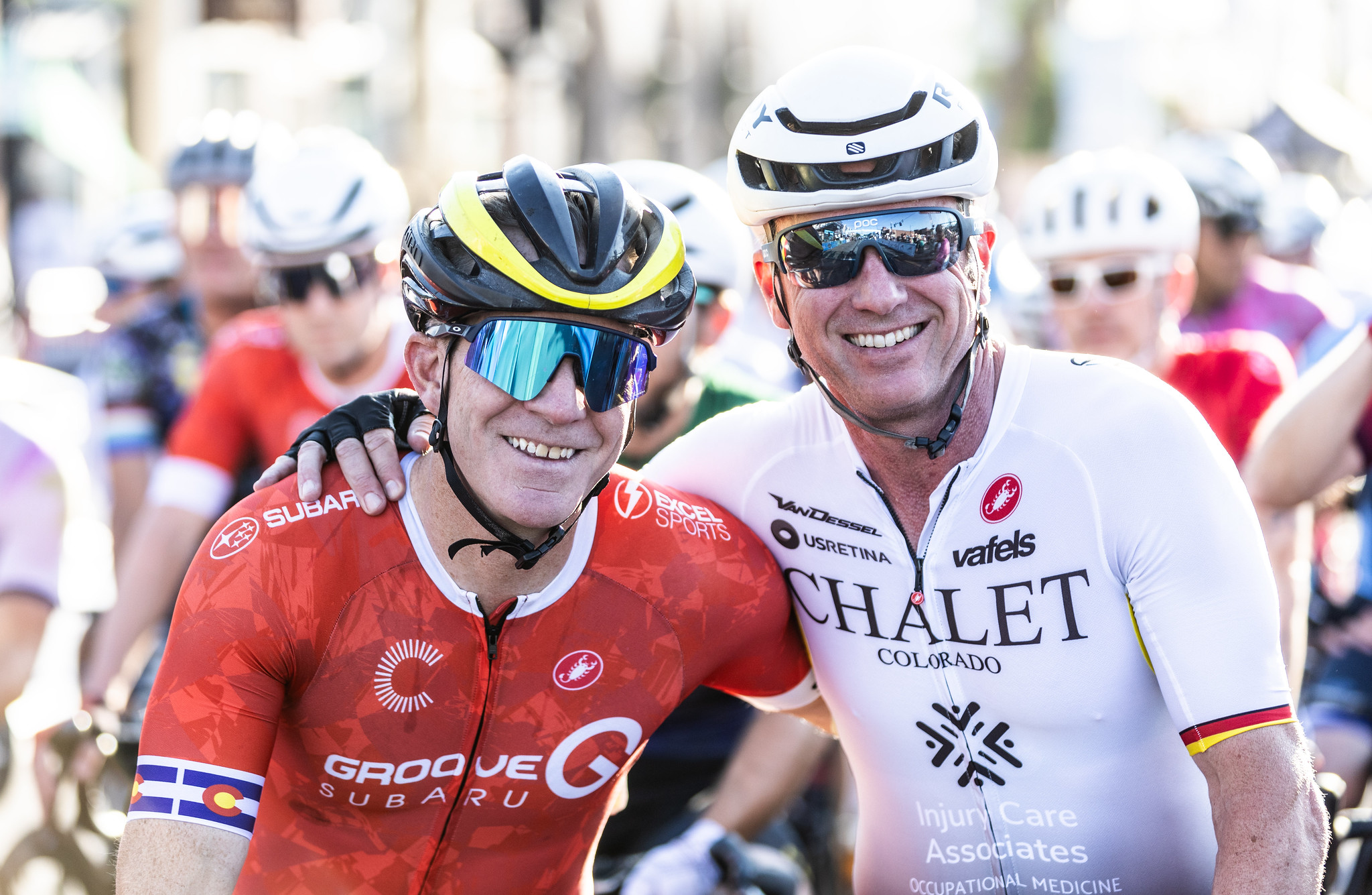 Two smiling cyclists in helmets and sunglasses pose together before a race, with others in the background.