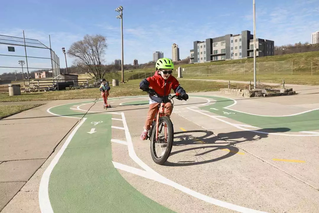 Two children ride bicycles on a paved track in a park on a sunny day, wearing helmets.