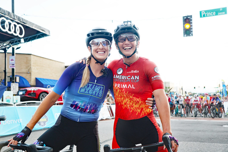 Two smiling cyclists in helmets and sunglasses pose together before a race, with others in the background.