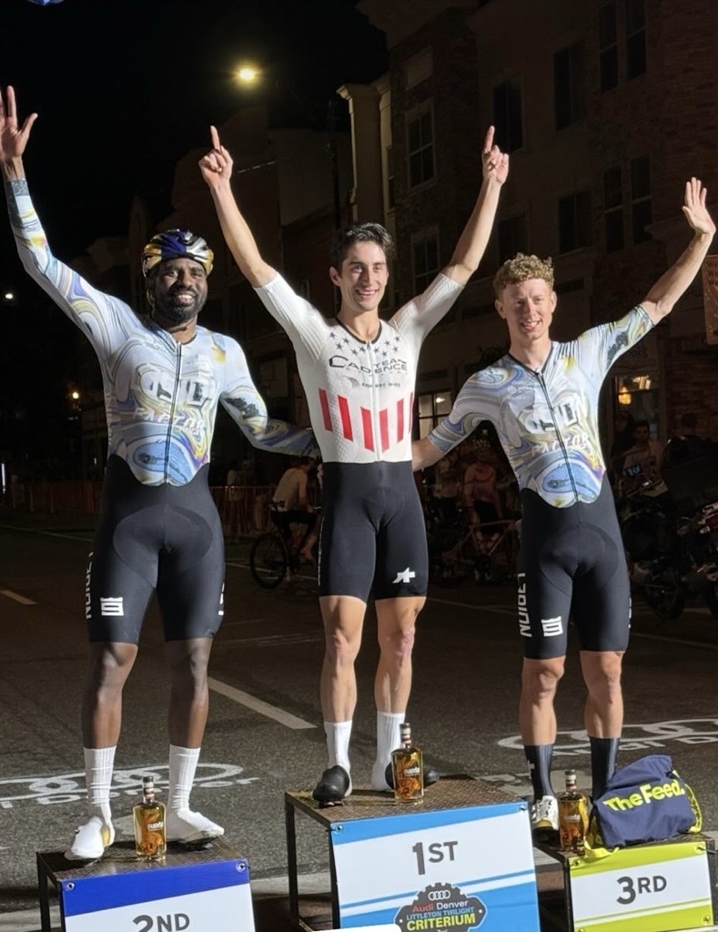 Three cyclists stand on podiums outdoors at night, celebrating their first, second, and third places.